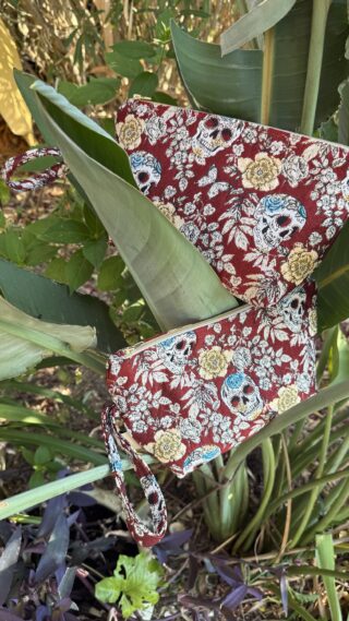 Red fabric bag with skull and floral pattern resting among green leaves and plant stems.