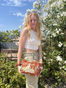 Young woman with blonde hair in a white crop top, beige pants, holding an orange patterned bag, standing in front of white flowering bushes under a blue sky.