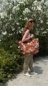 Woman standing outdoors with a large brown patterned tote bag over her shoulder, in front of a flowering shrub with white blossoms.