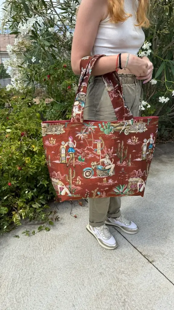 Person standing outdoors holding a large red tote bag with retro Route 66 desert illustrations, wearing a white tank top and khaki pants.
