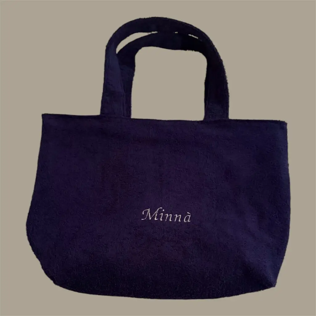 Navy tote bag with two handles on a beige background; script text 'Minna' embroidered on front