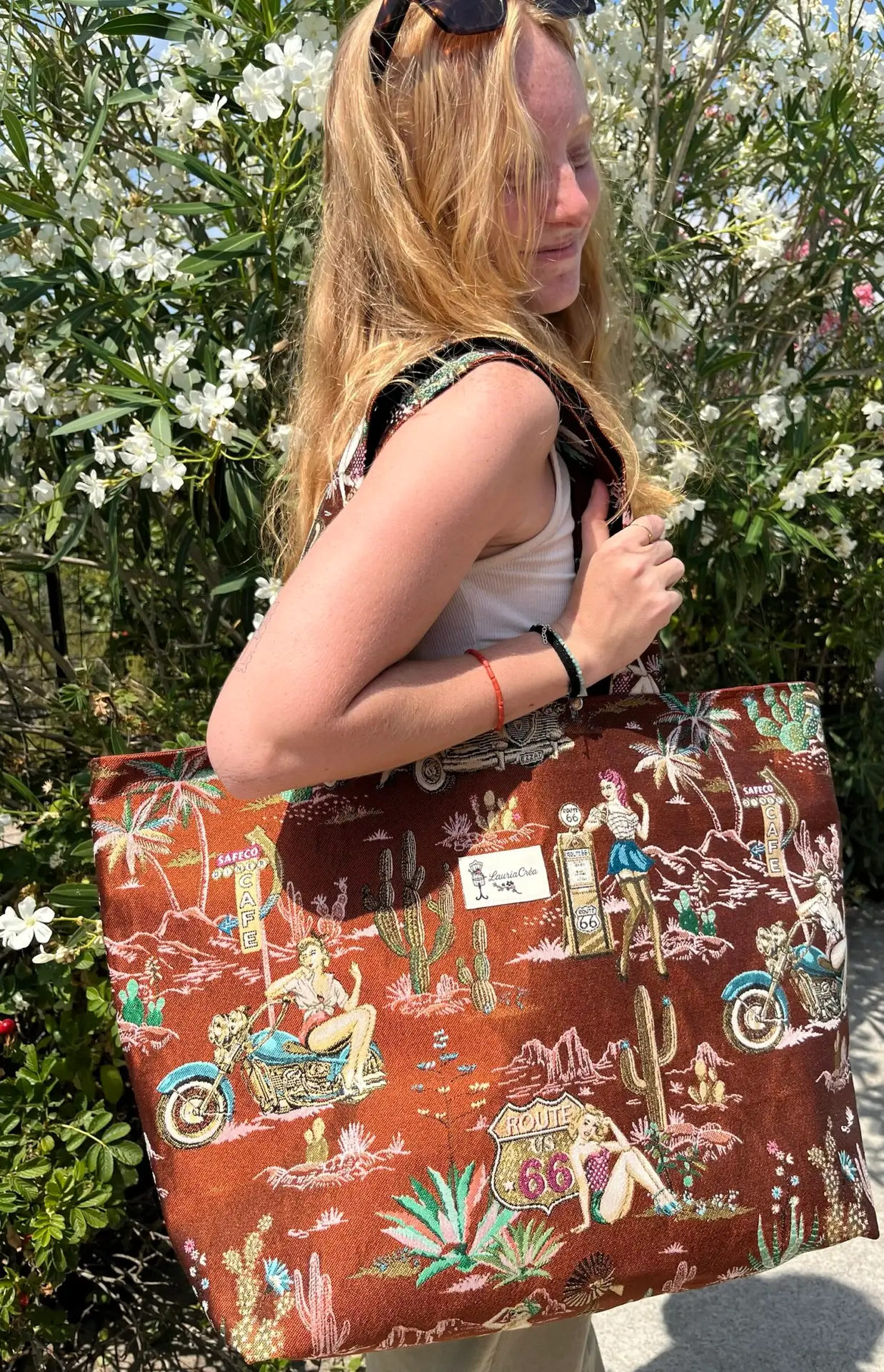 Woman with long blonde hair and sunglasses on her head, wearing a white tank top, carrying a large red-brown tote bag with a vintage Route 66 design among white flowering bushes in sunlight.