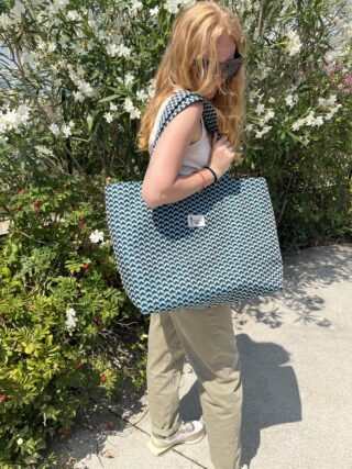 Woman with long red hair wearing sunglasses carries a large blue geometric tote bag outdoors, white flowers in the background.