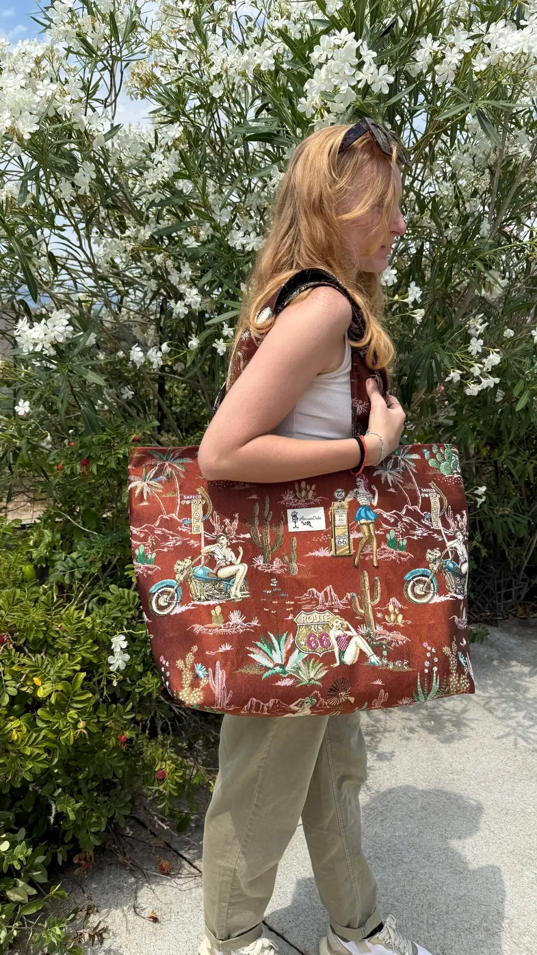 Profile view of a woman in a white tank top and khaki pants, carrying a large brown tote bag with vintage Route 66 graphics amid white flowering shrubs.