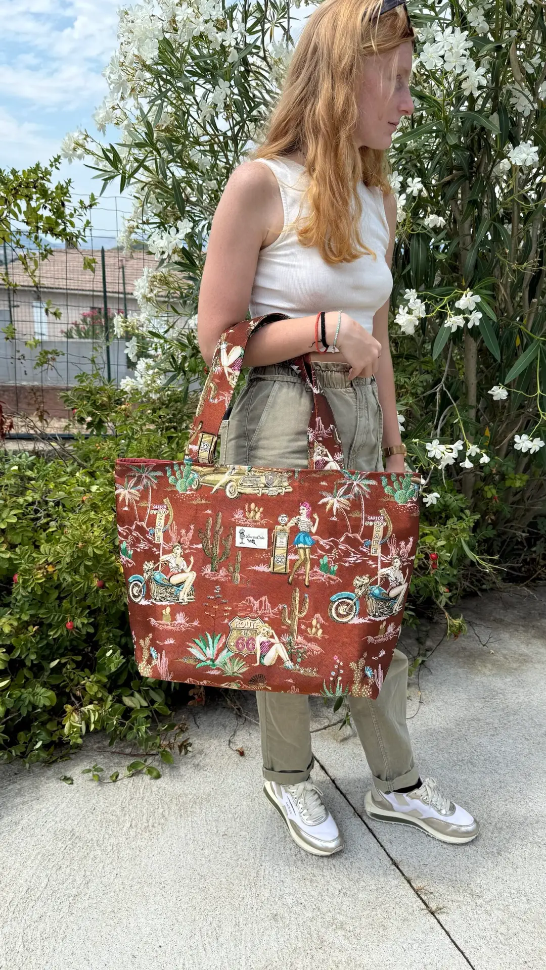 Side view of a person in a white sleeveless top and khaki pants holding a large red-brown tote bag with vintage travel illustrations in a garden setting.