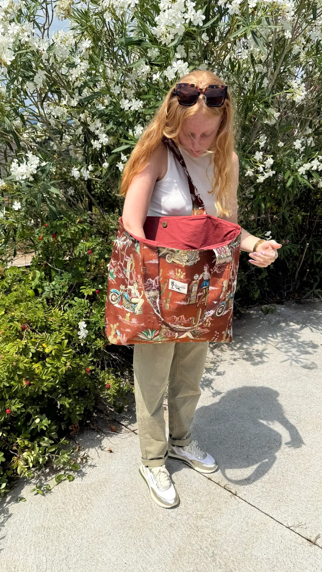 Blond woman in a white tank top and khaki pants looks into a large red-pattern tote bag outdoors, sunglasses on her head, with white flowering bushes in the background.